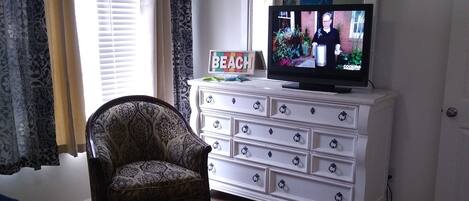 Una habitación con una cama, un televisor sobre una cómoda, una silla y un ventanal con cortinas.