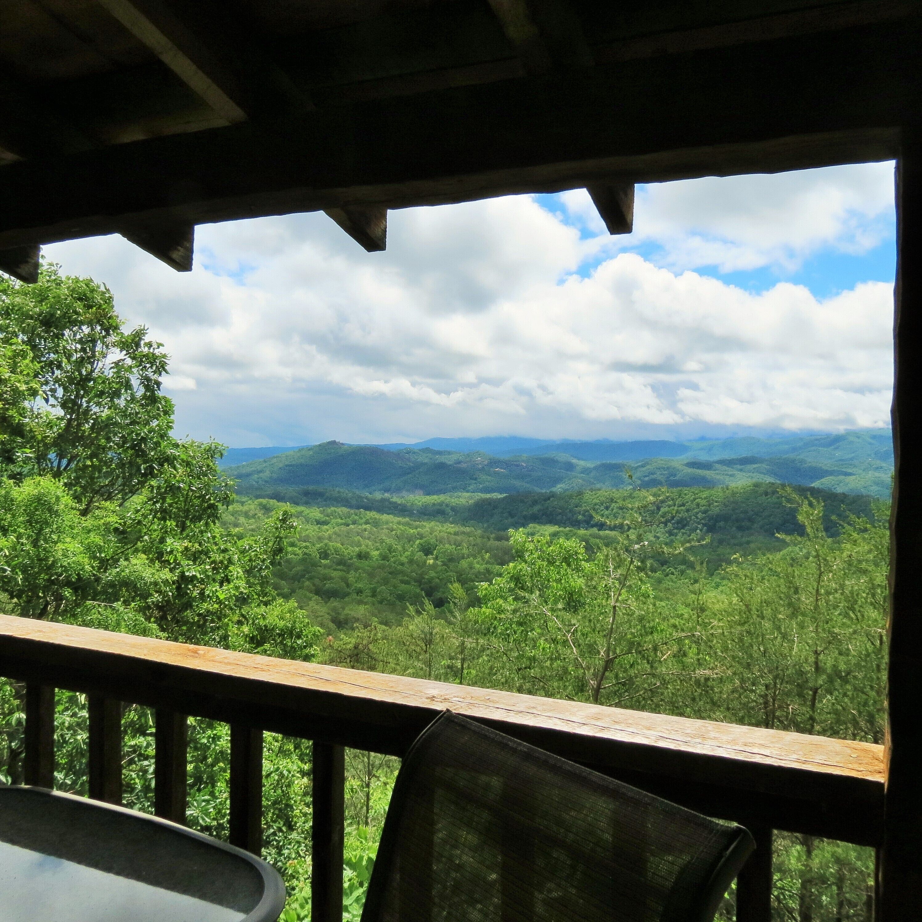 A view from a covered outdoor area with a wooden railing, overlooking a lush green landscape with distant hills and a partly cloudy sky.