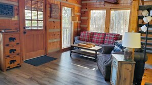A cozy cabin interior with a wooden door, a sofa with plaid cushions, a small table with a tray, and a lamp on a wooden side table.
