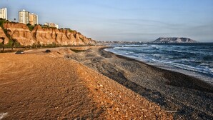 Een strand met een kiezelstrand, een klif met gebouwen en een berg in de verte.