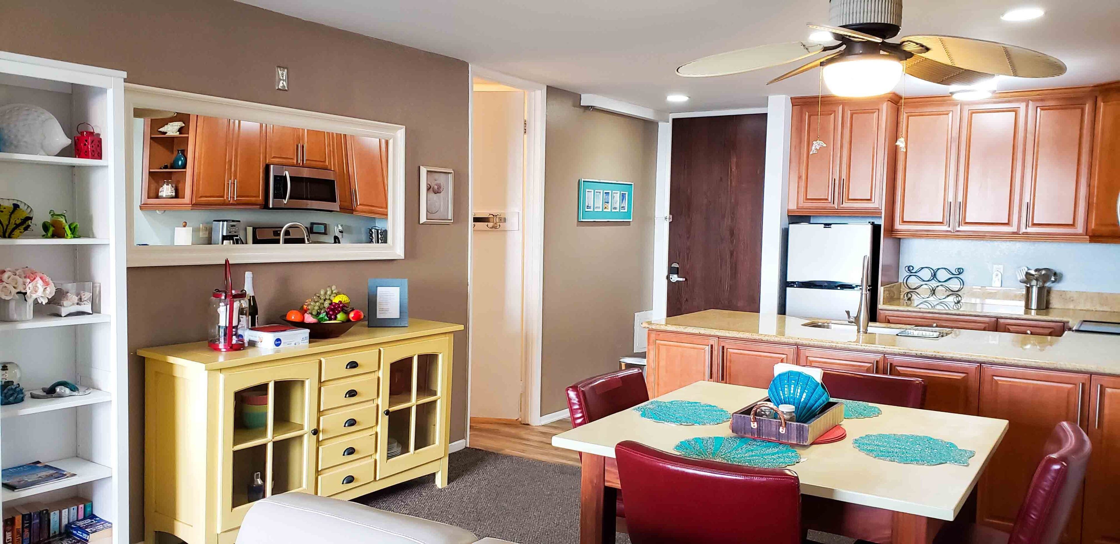 Una cocina compacta con armarios de madera, un microondas y un refrigerador. Hay una mesa de comedor con sillas y un ventilador de techo.