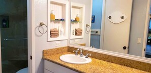 A bathroom with a granite countertop, a white sink, and a mirror above it. There are shelves with toiletries and towels. - STRIKING OCEAN VIEW STUDIO D210 FACING OCEANSIDE PIER (Oceanside)
