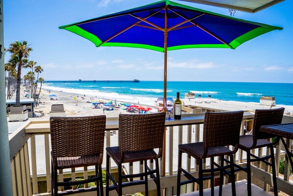 A balcony with wicker chairs and a large umbrella overlooking a beach with a pier in the distance. - STRIKING OCEAN VIEW STUDIO D210 FACING OCEANSIDE PIER (Oceanside)