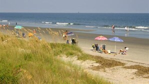 Ein Strand mit Menschen, die unter Sonnenschirmen entspannen, grasbewachsenen Dünen und dem Meer im Hintergrund.