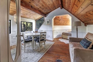 A cozy loft-style room with a wooden ceiling, a dining area with a table and chairs, a sofa, and a window providing natural light.
