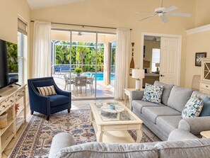 A spacious living room with a television, a sofa, a coffee table, and a chair. There is a sliding glass door leading to a pool area with outdoor furniture. - Naples Luxury Spotless 4 Bdrm Heated Pool Walk Bike To Beach (Naples)