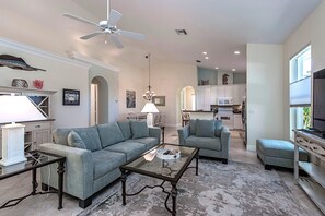 A spacious living room with a sofa, armchair, and coffee table, a ceiling fan, and a kitchen area in the background. - Naples Luxury Spotless 4 Bdrm Heated Pool Walk Bike To Beach  (Naples)