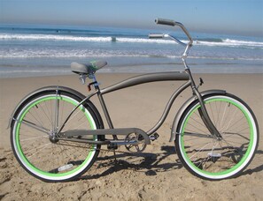 Een fiets met een groen en wit frame, een zwarte zadel en stuur, geparkeerd op een zandstrand met de oceaan op de achtergrond.