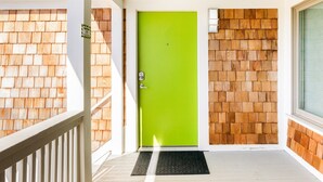 Una puerta verde brillante con felpudo, paredes de madera y una barandilla blanca.