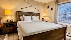 A bedroom with a wooden bed, two bedside tables with lamps, a ceiling fan, a window with a view, and a wall-mounted clock.