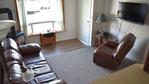 A living room with a brown leather sofa, a brown leather armchair, a small wooden side table, a lamp, and a flat-screen TV mounted on the wall. - 1 Block From The Beach! (Seward)