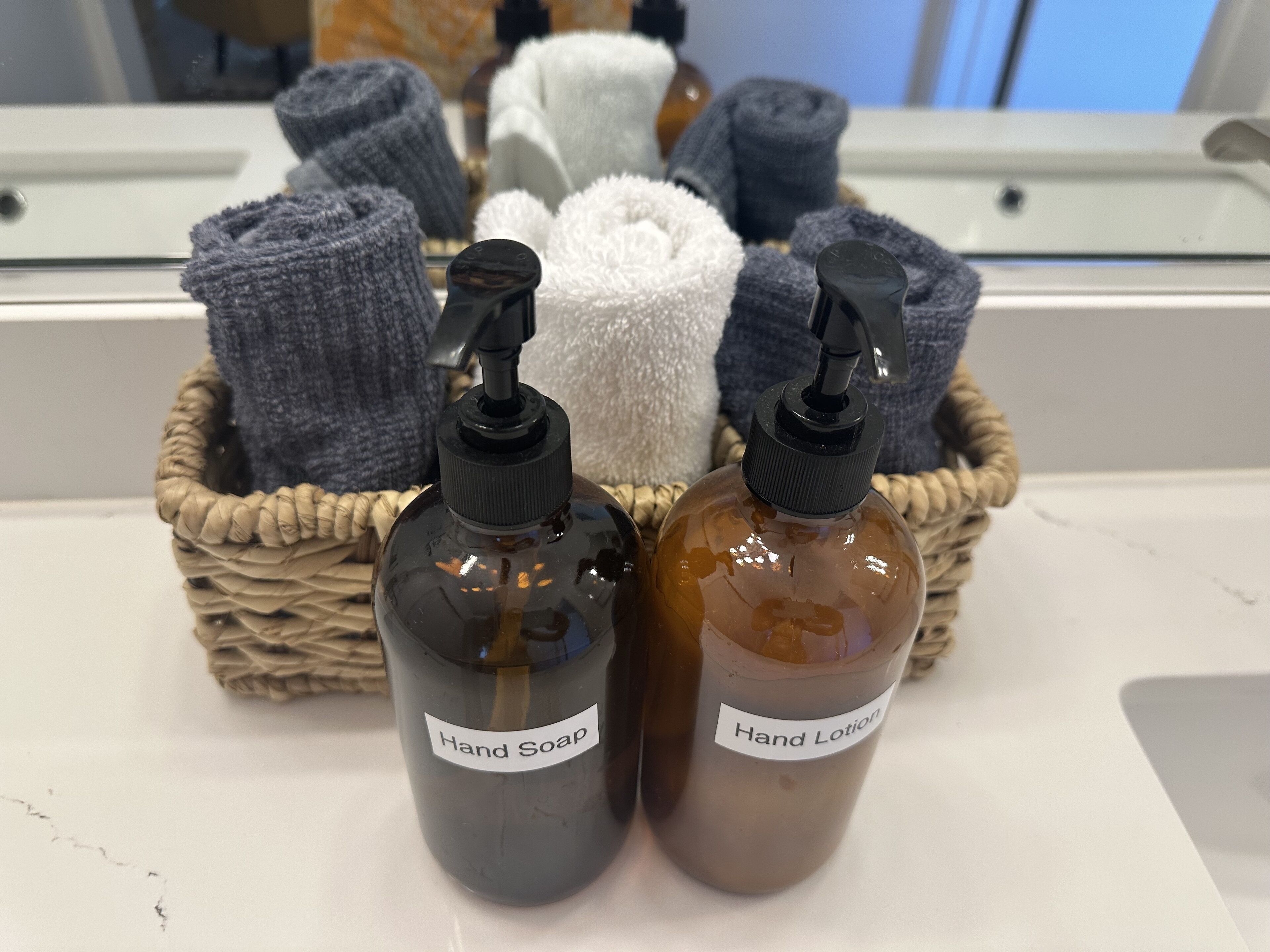 Deux bouteilles de savon liquide et de lotion pour les mains dans un panier en osier, accompagnées de serviettes enroulées.
