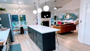 A modern kitchen with a large island, dining area with chairs, and a living room with a sofa and fireplace. - Emerald House - striking, remodel in tranquil, central neighborhood (Redding)