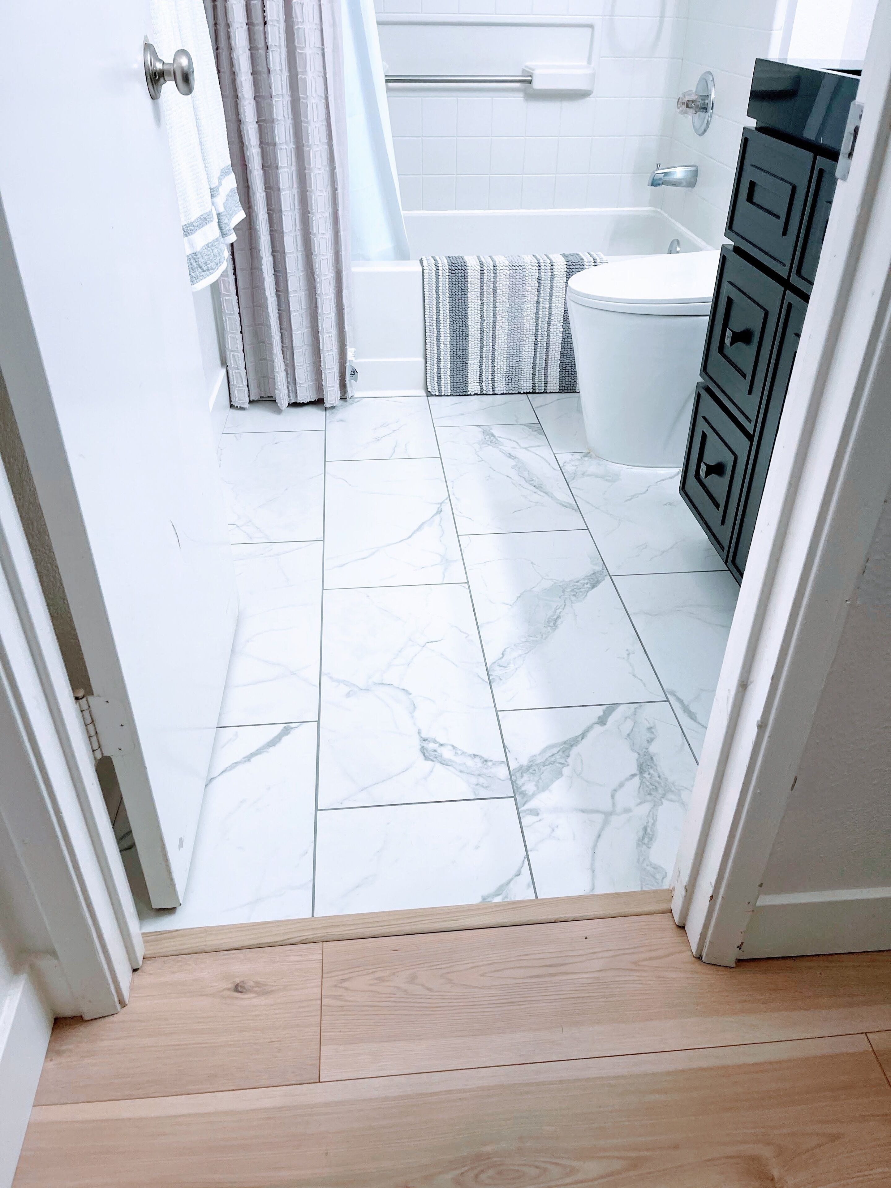 A bathroom with white marble tiles, a bathtub, a toilet, and a wooden floor leading to a door.