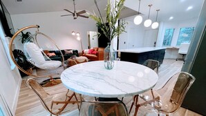 A modern living room with a round marble table, clear chairs, a hanging chair, a sofa, and a kitchen in the background. - Emerald House - striking, remodel in tranquil, central neighborhood (Redding)