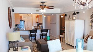 A living room with a sofa, a coffee table, a lamp, and a dining table with a candle. There is a kitchen in the background with cabinets and a refrigerator.