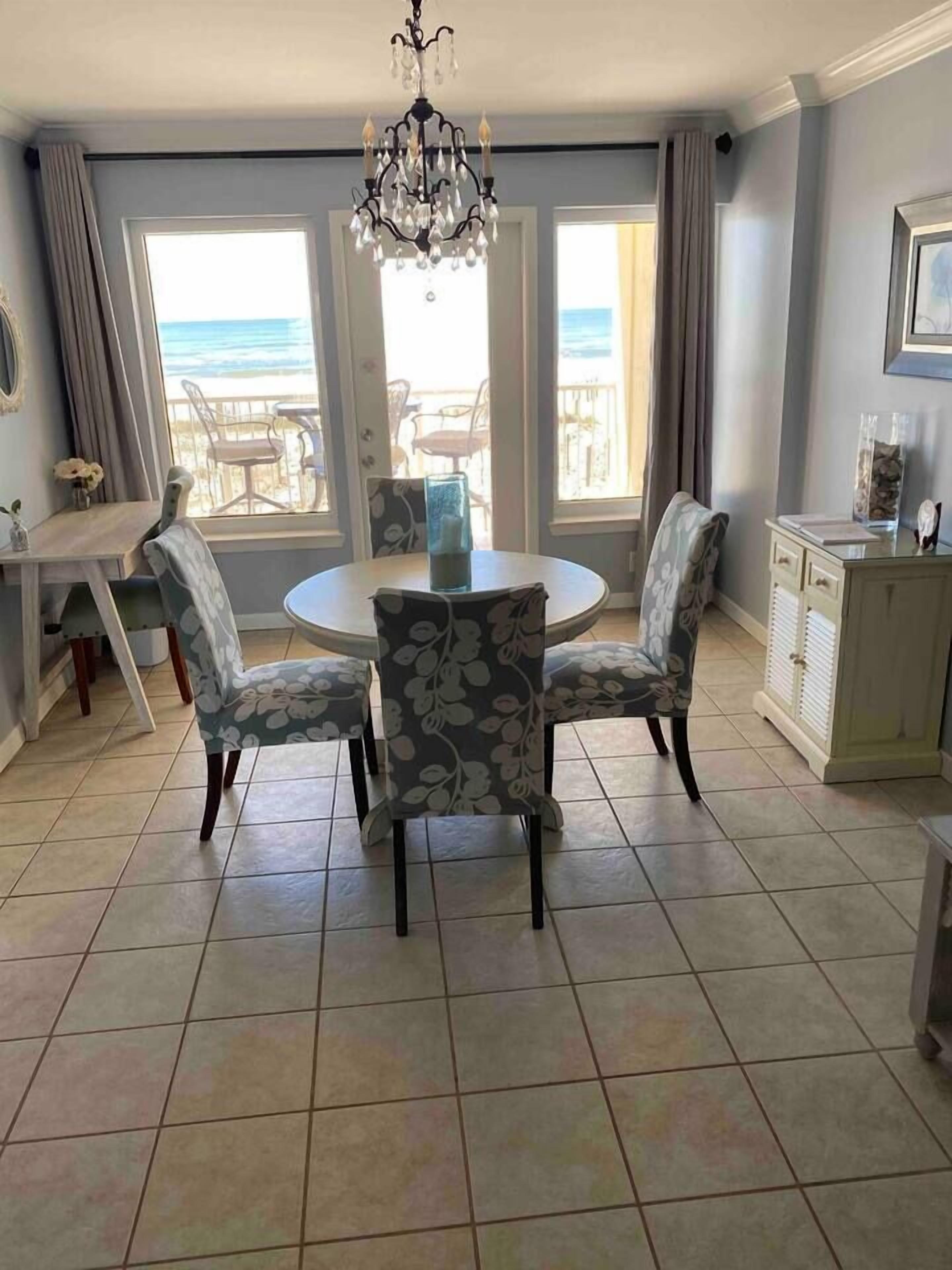A dining area with a round table and floral chairs, a chandelier, and a view of the beach through large windows.