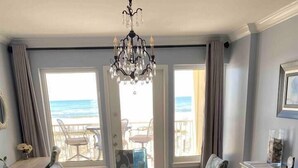 A dining area with a round table and floral chairs, a chandelier, and a view of the beach through large windows.