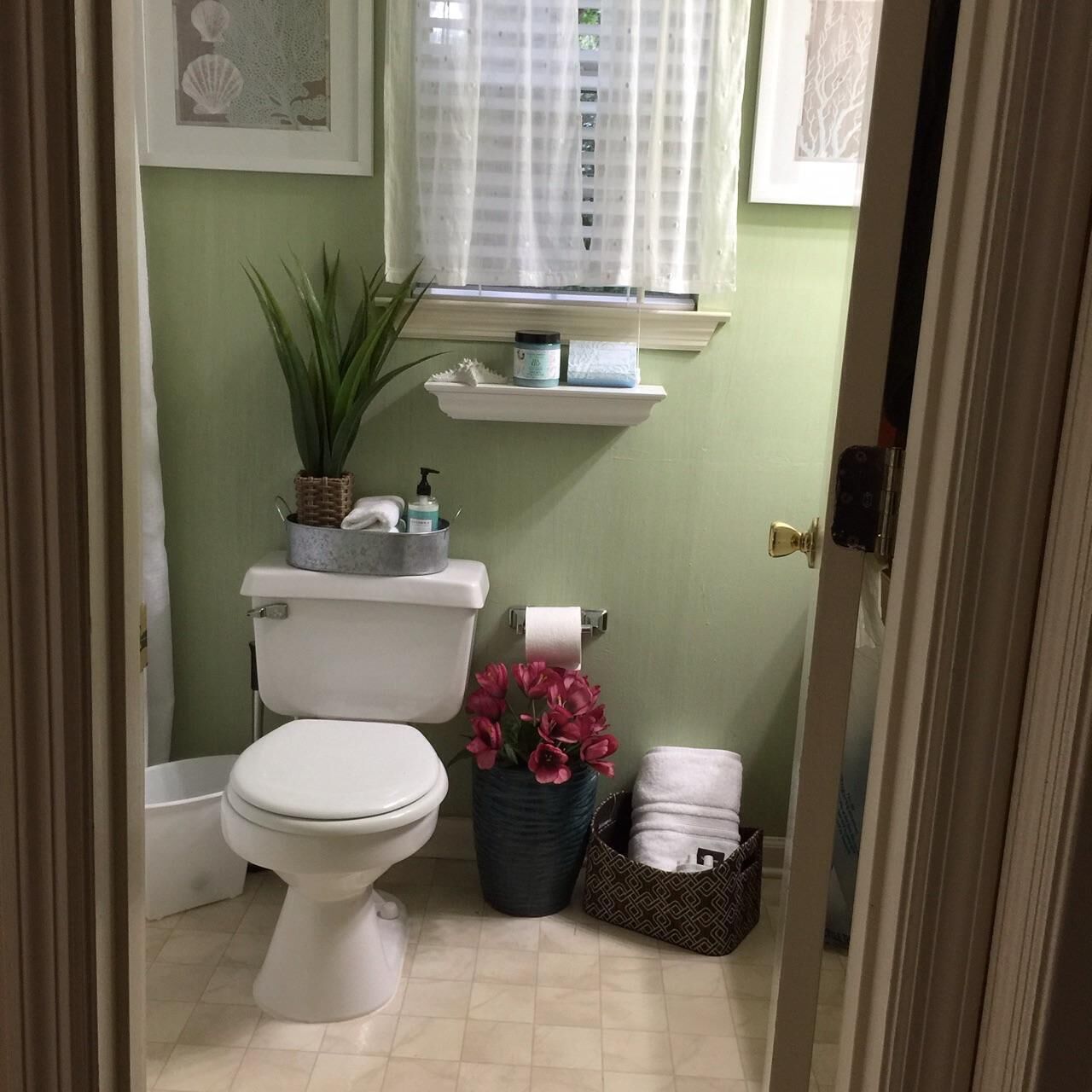 A compact bathroom with a toilet, a sink, a window with sheer curtains, and a shelf with toiletries.