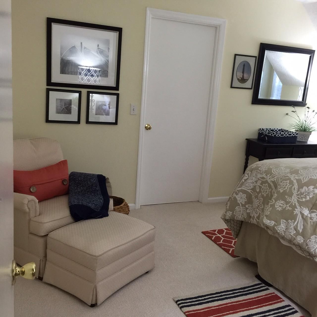 A bedroom with a bed, a chair, a small table, and a door. There are framed pictures on the wall and a plant on the table.