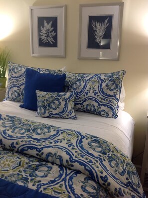 Een bed met beddengoed met patroon, een blauw kussen en twee ingelijste botanische prints aan de muur.
