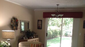 Un salon avec un canapé beige, une table basse sur laquelle se trouvent un vase et un panier de fruits, une lampe et une grande fenêtre donnant sur un jardin.