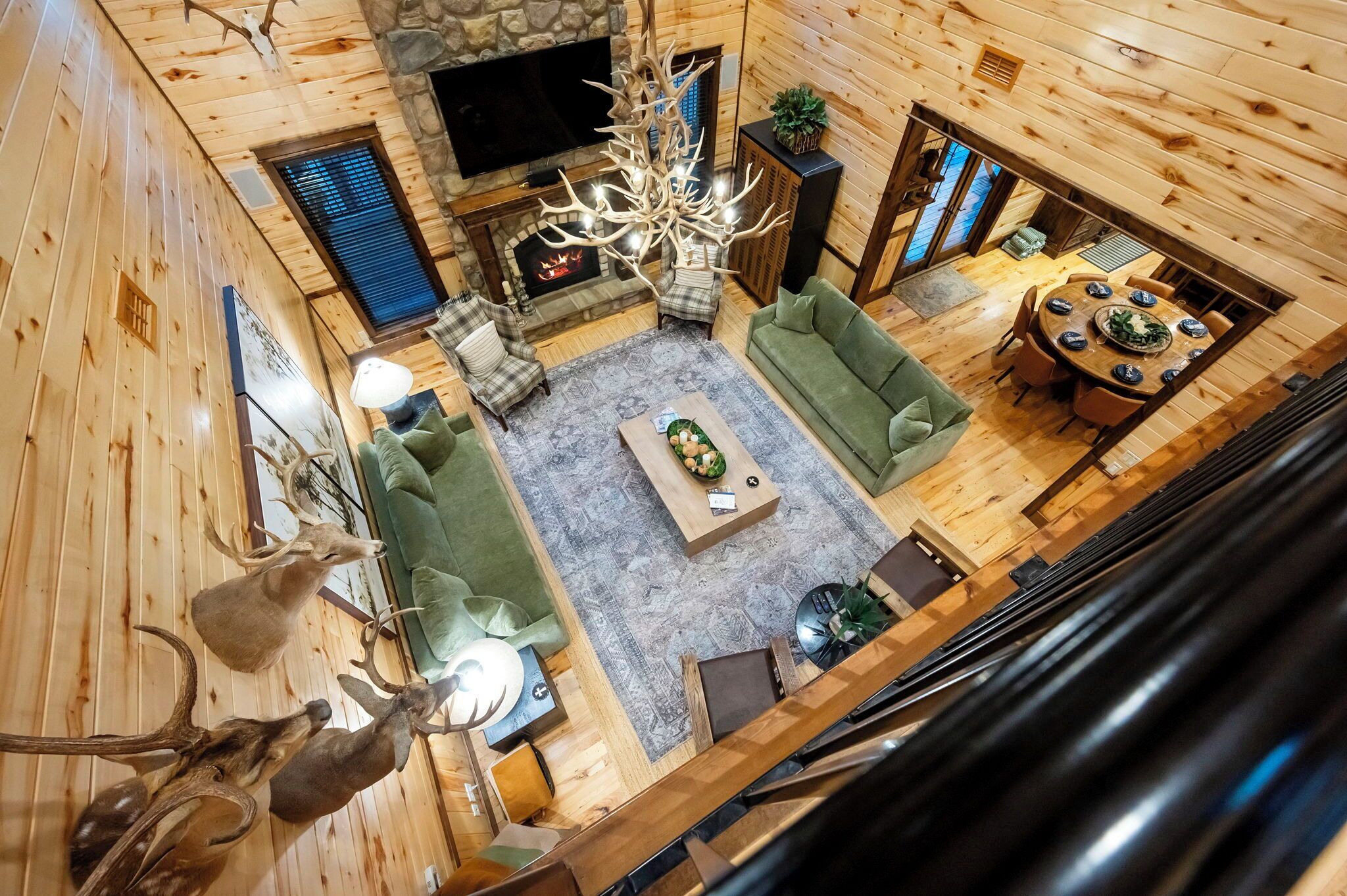 A wooden cabin interior with a fireplace, green sofas, a dining table, and a chandelier.