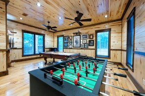 Ett rum med ett fotbollsspel, takfläktar, inramade tavlor och stora fönster.