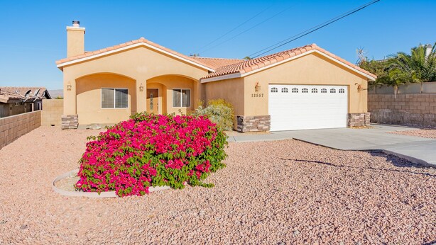 Una casa de un solo piso con fachada color beige, puerta de garaje blanca y un pequeño jardín con flores rosadas.