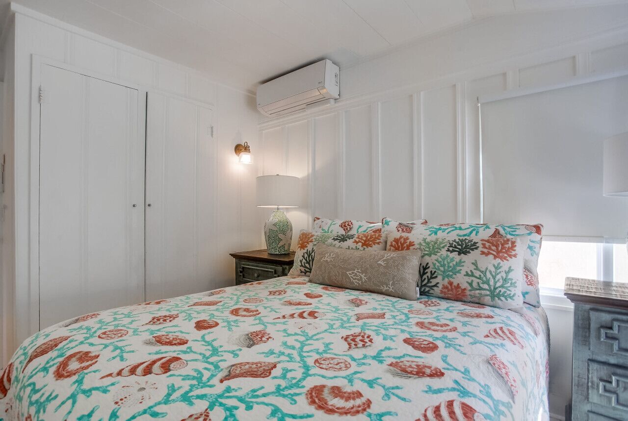 Een slaapkamer met een bed, witte wanden, een nachtkastje, een lamp en een airconditioning.
