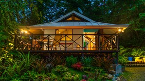 A two-story wooden structure with a balcony, surrounded by lush vegetation and a well-maintained garden.