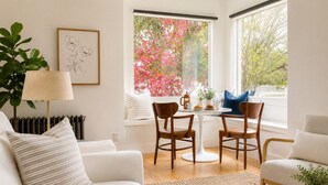 Ein modernes Wohnzimmer mit einem weißen Sofa, einem Couchtisch und zwei Sesseln. Ein Heizkörper, eine Lampe und ein Fenster mit Blick auf rosa Blumen sind ebenfalls zu sehen.