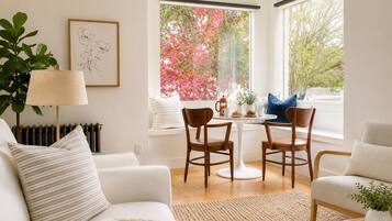Ein modernes Wohnzimmer mit einem weißen Sofa, einem Couchtisch und zwei Sesseln. Ein Heizkörper, eine Lampe und ein Fenster mit Blick auf rosa Blumen sind ebenfalls zu sehen.