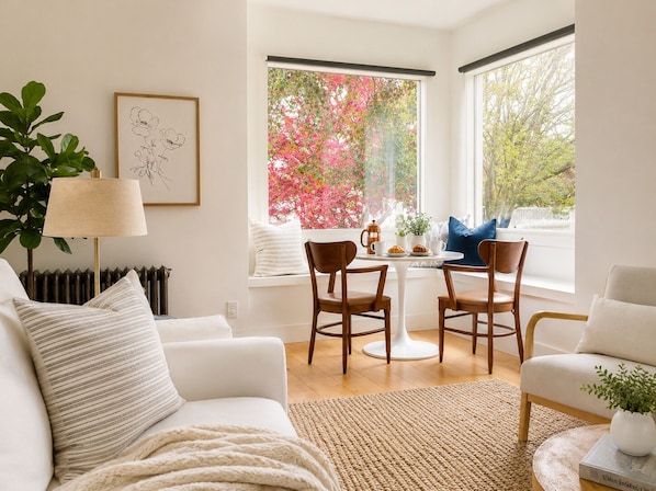 A modern living room with a white sofa, a coffee table, and two chairs. There is a radiator, a lamp, and a window with a view of pink flowers.