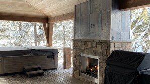 A wooden deck with a stone fireplace and a grill, surrounded by trees.