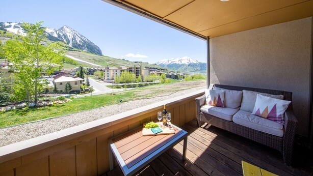 A balcony with a view of a mountain range, a wicker sofa, and a small table with a bottle and glasses. - Rustic Modern Retreat! 3bed/2bath Close to Lifts (Crested Butte)