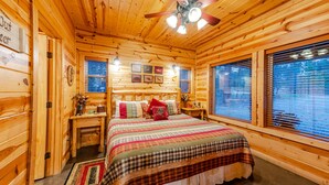 Un dormitori de cabana de fusta amb un llit, parets de fusta, un ventilador de sostre i una finestra amb persianes.