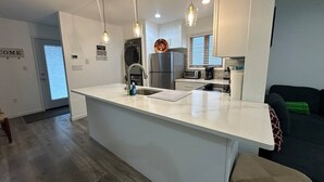 Een moderne keuken met een centraal eiland, roestvrijstalen apparatuur, hanglampen en uitzicht op de woonkamer.