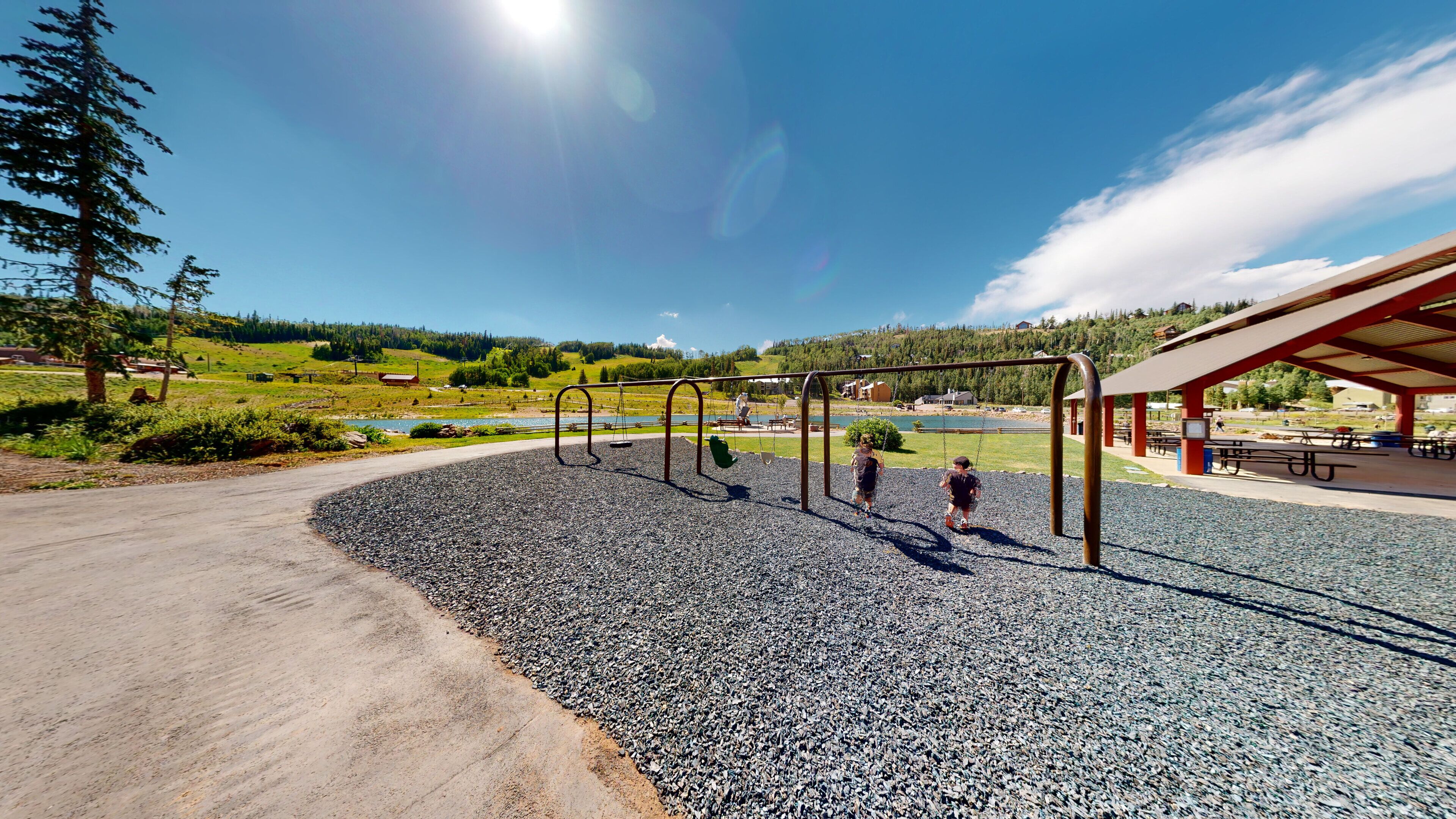 A playground with swings and a covered picnic area, surrounded by a gravel path and a scenic view of a lake and hills.