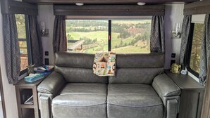 Ein gemütliches Wohnmobil-Interieur mit einer Ledersitzgruppe, einem kleinen Tisch mit Lampe und Blick auf eine reizvolle Landschaft durch die Fenster.