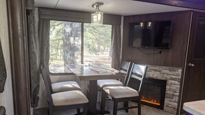 Ein modernes Wohnzimmer mit Kamin, Esstisch mit Stühlen, Wand-Fernseher und großem Fenster mit Vorhängen.