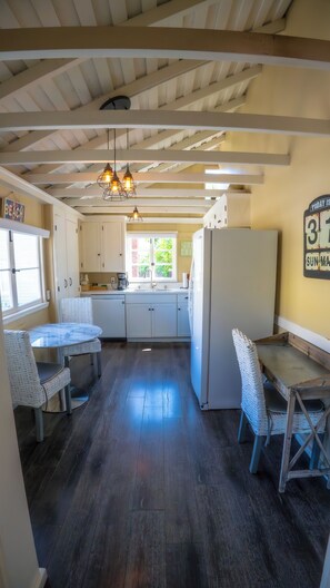 Una cocina con armarios blancos, un refrigerador, una mesa de comedor con sillas y una lámpara colgante.