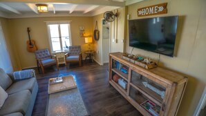 A living room with a couch, two chairs, a television, a guitar, a lamp, and a bookshelf. - Green Beach House 500' to the Waves- Electric Vehicle Charger! (Laguna Beach)