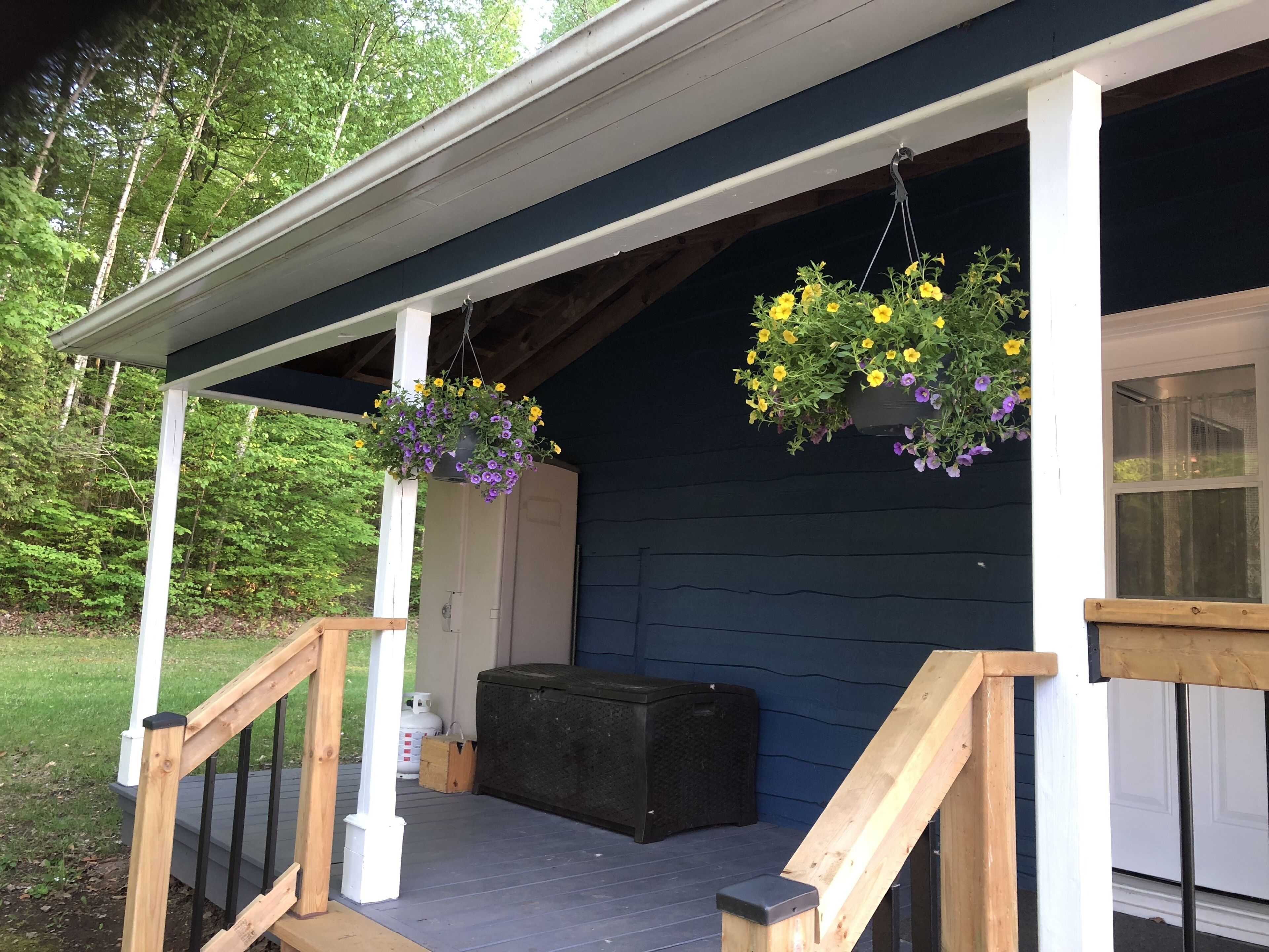 Une véranda couverte avec une balustrade en bois, un coffre de rangement noir et des jardinières suspendues.