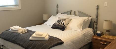 Una cama bien hecha con sábanas blancas, un edredón gris oscuro y toallas dobladas. Hay una mesita de noche con una lámpara, y una mesita con una lámpara, además de un cuadro en la pared.