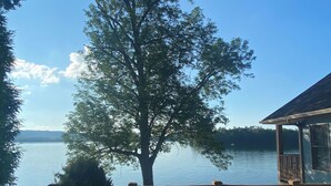 A wooden deck with a railing overlooking a calm lake, a large tree in the center, and a building with a balcony to the right. - Luxurious Lakefront Living With Amenities close to Cooperstown (Richfield Springs)