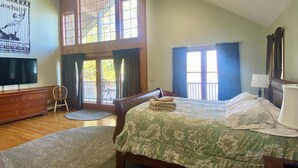 A spacious bedroom with a large bed, a ceiling fan, a TV, and a view of the outdoors through a large window. - Luxurious Lakefront Living With Amenities close to Cooperstown (Richfield Springs)
