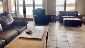 A living room with a checkered coffee table, leather sofas, and a view of a lake through large windows. - Luxurious Lakefront Living With Amenities close to Cooperstown (Richfield Springs)
