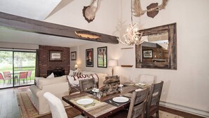A dining area with a rustic wooden table, chairs, and a chandelier. There is a fireplace with a brick wall and a large mirror.