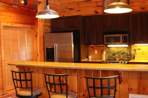A kitchen with wooden cabinets, a stainless steel refrigerator, a microwave, and a bar area with chairs.